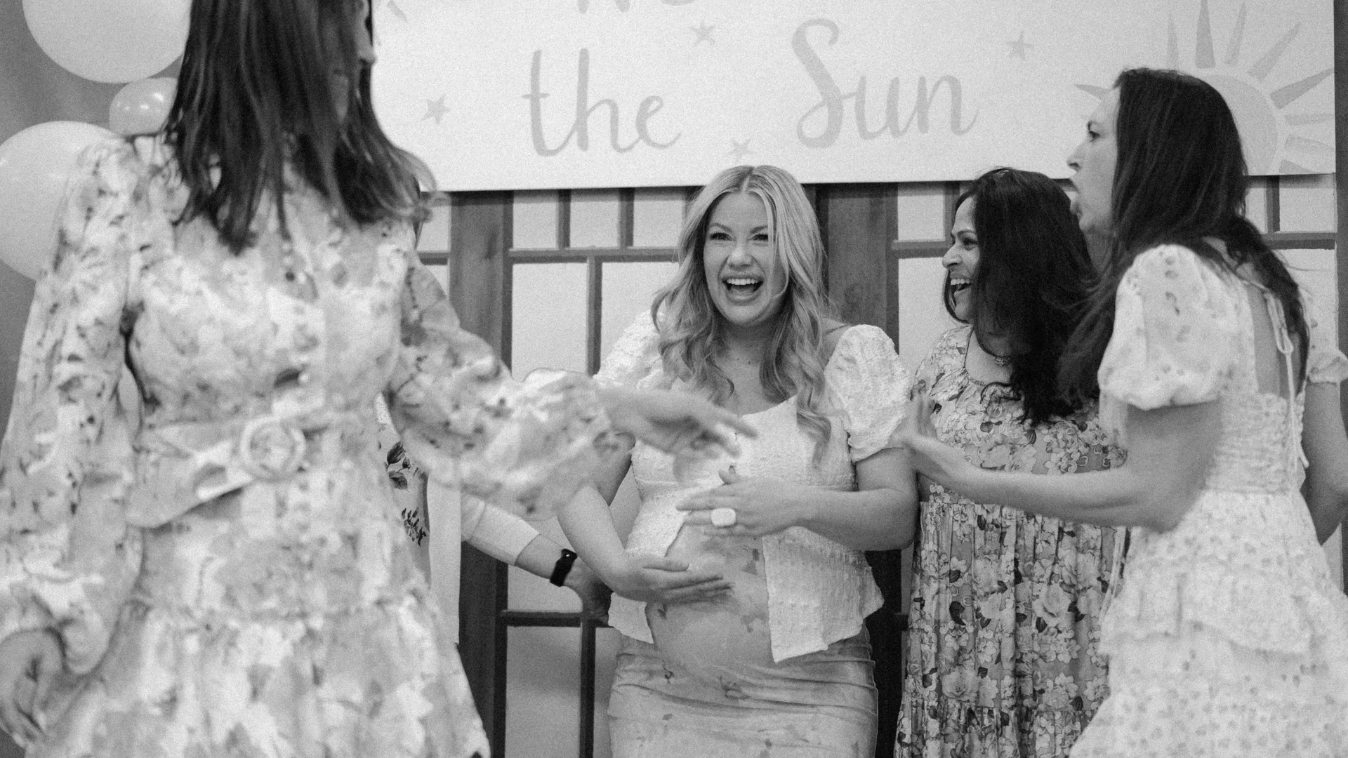Mum-to-be laughing with friends in front of a Here Comes the Sun banner at her baby shower